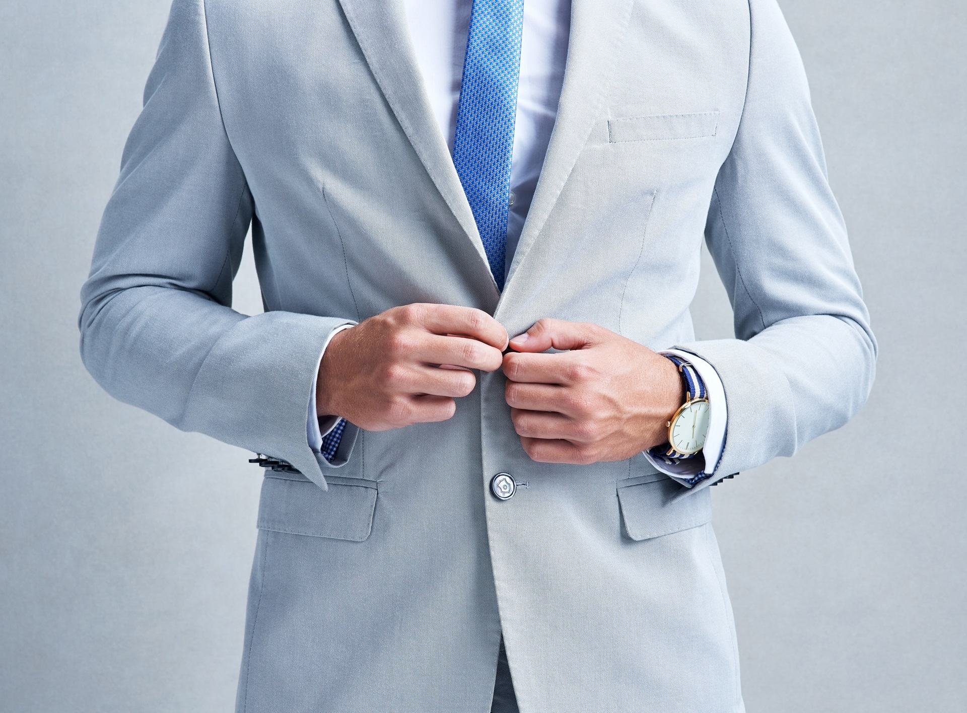 A person in a light gray suit and blue tie buttons their jacket. They wear a wristwatch and shirt cuffs are visible. A person in a light gray suit and blue tie buttons their jacket. They wear a wristwatch and shirt cuffs are visible.