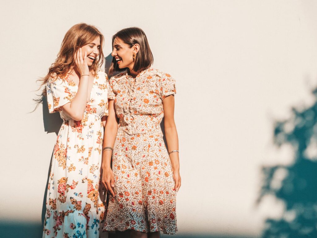 Two women in floral dresses stand against a light-colored wall, smiling and looking at each other in bright sunlight.