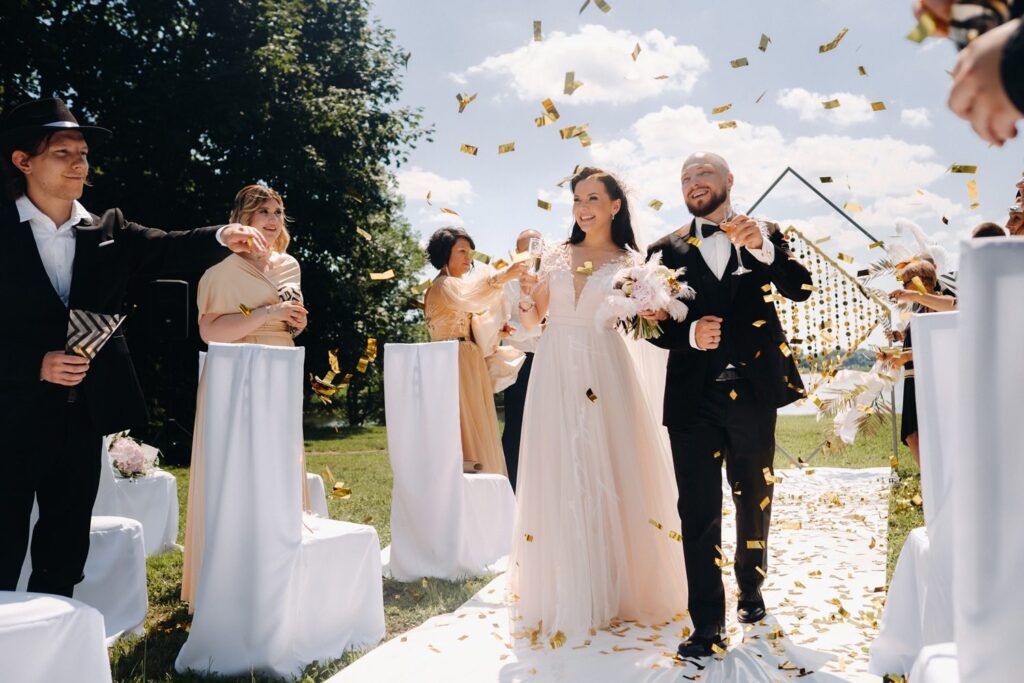 A bride and groom walk down an outdoor aisle while guests throw gold confetti to celebrate their wedding. The sun is shining and everyone is smiling.
