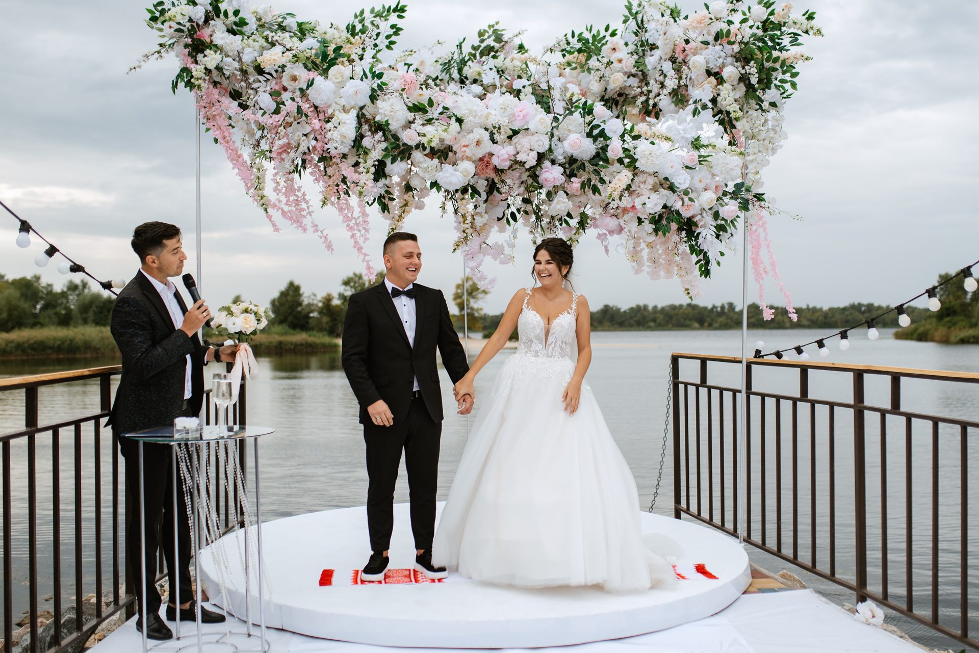 A bride and groom stand holding hands on an outdoor platform decorated with flowers, while an officiant speaks into a microphone beside them near a waterfront.