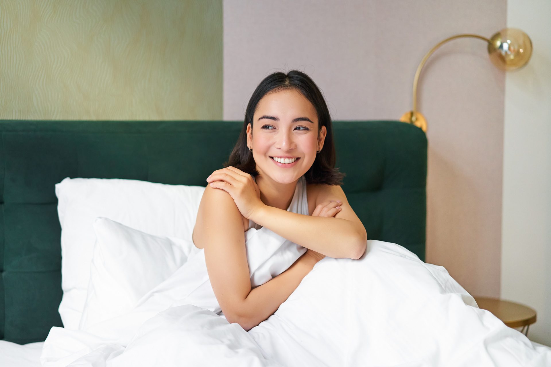 A woman sits on a bed covered in white sheets, smiling and looking to the side. A green headboard and gold lamp are visible in the background.