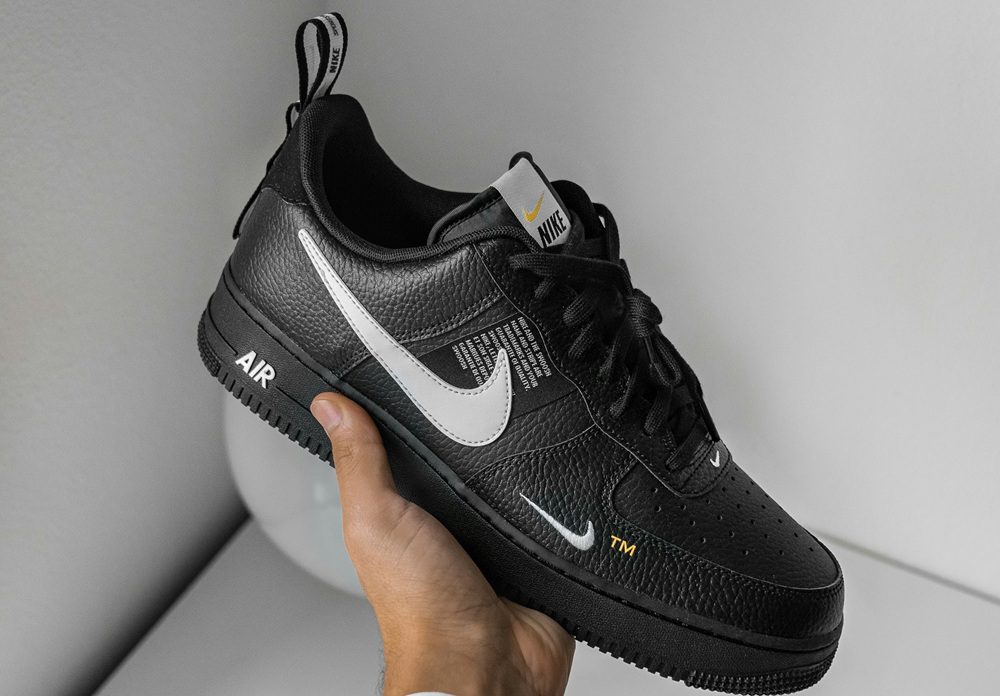 A hand holds a black Nike Air Force 1 sneaker with white swoosh logos and "AIR" text on the black sole, against a plain background.