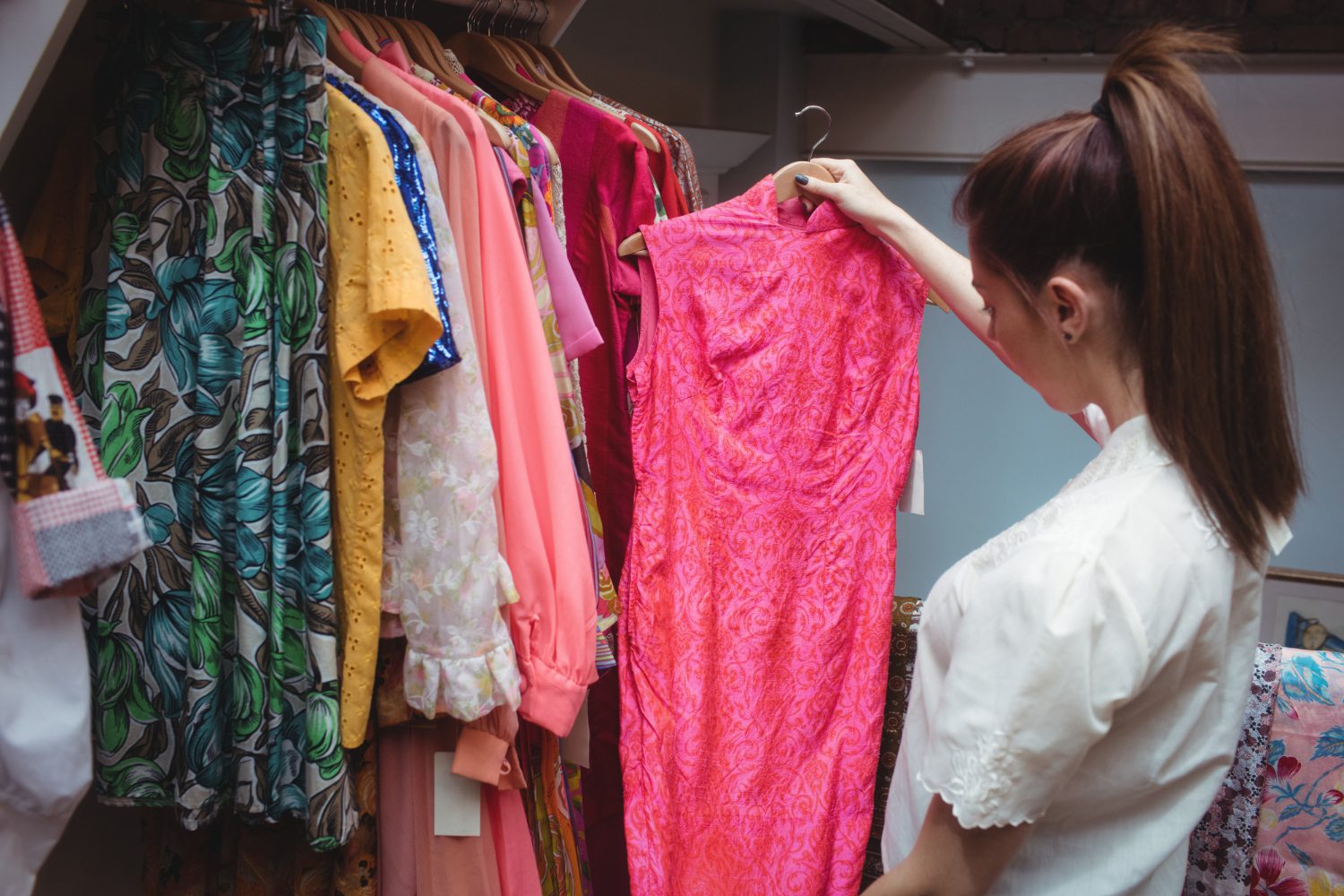 A woman examines a bright pink dress on a hanger in front of a clothing rack filled with various colorful garments.