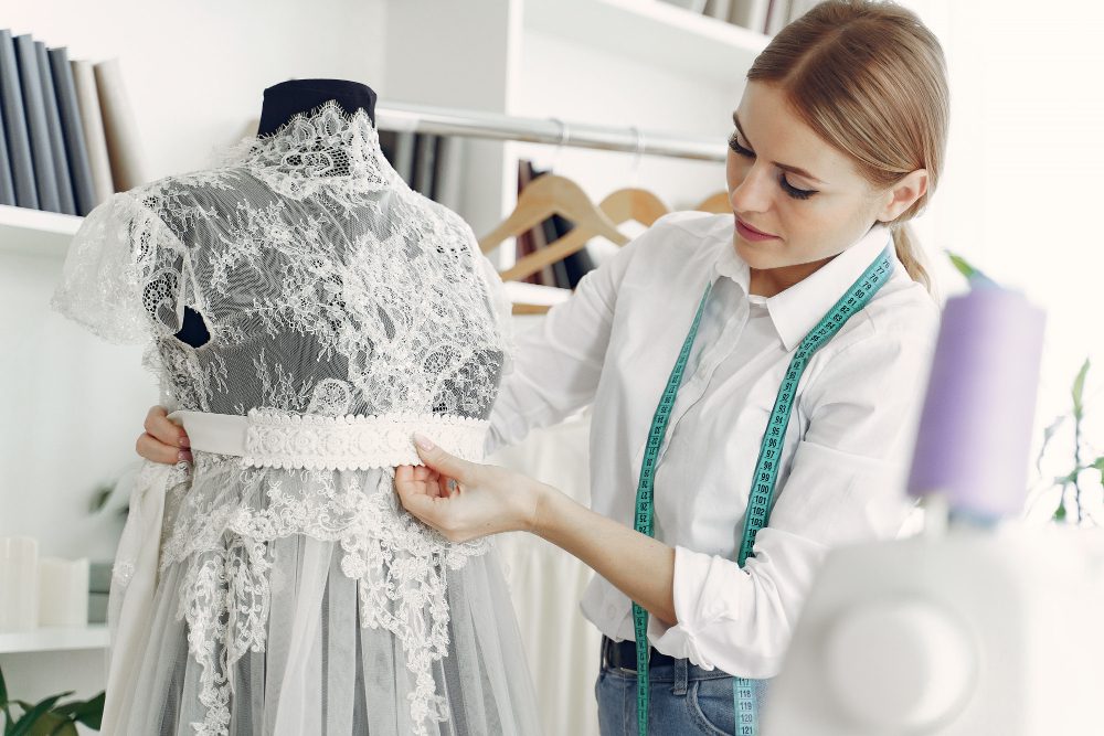 A woman adjusts a lace wedding dress on a mannequin, with a measuring tape around her neck in a sewing studio.