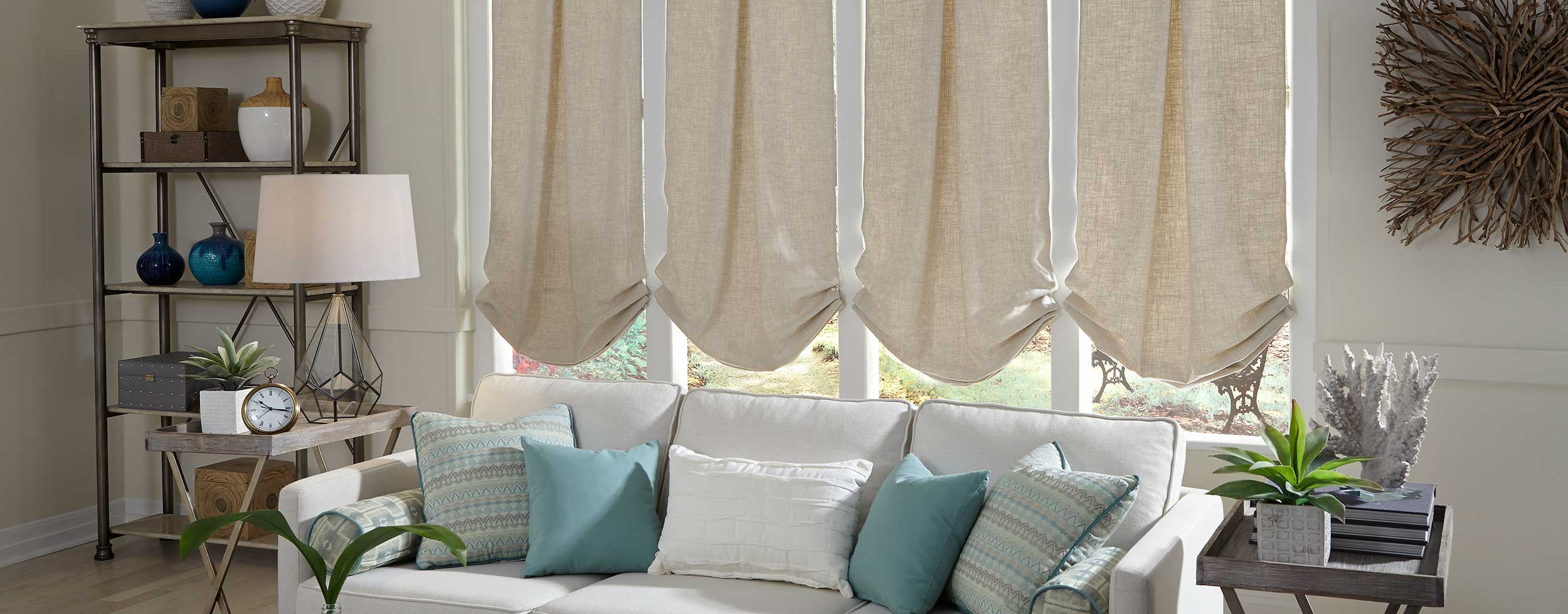 A white sofa with blue and patterned pillows sits in front of windows with beige Roman shades. A floor lamp, metal shelf, and decorative items are visible in the living room.