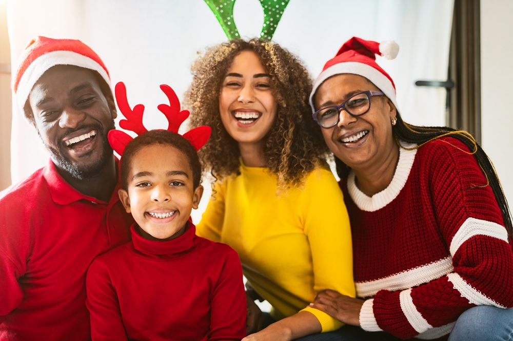 Four people wearing festive clothing, Santa hats, and reindeer antlers smile together indoors, suggesting a holiday celebration.