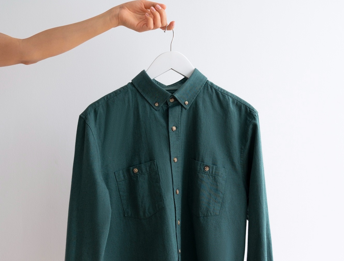 A hand holds a hanger with a green button-up shirt against a plain white background.