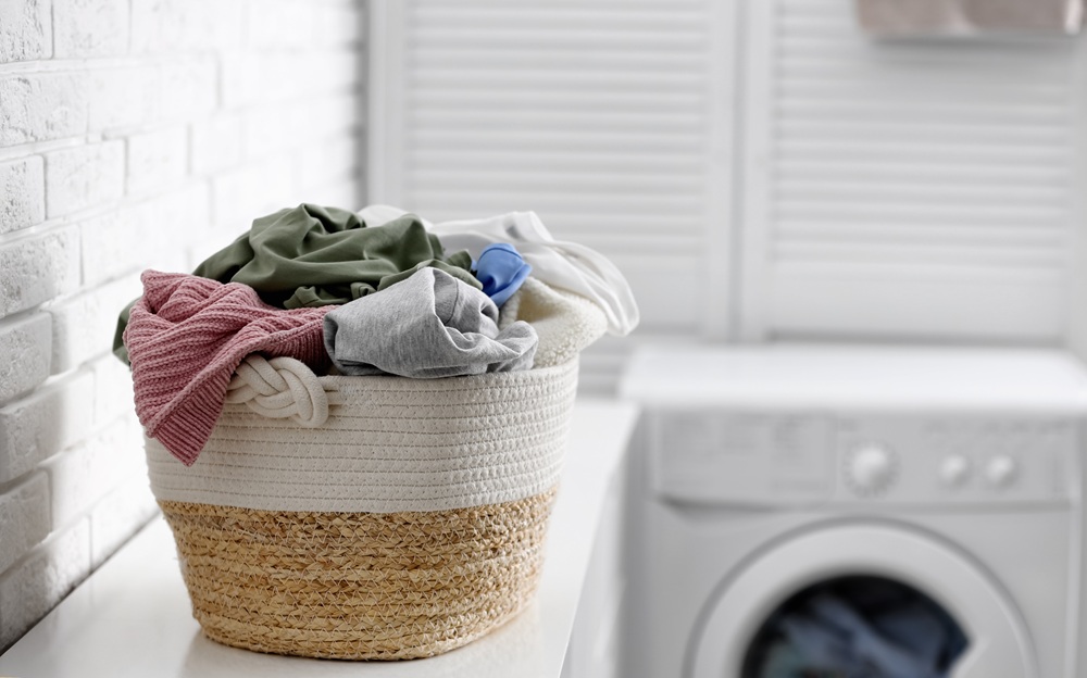 A woven laundry basket filled with assorted clothes sits on a counter next to a washing machine in a bright laundry room. A woven laundry basket filled with assorted clothes sits on a counter next to a washing machine in a bright laundry room.