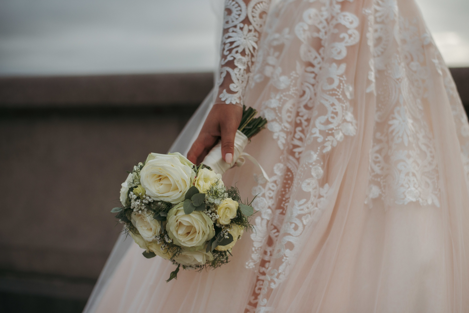 A bride in a lace wedding dress holds a small bouquet of white roses and greenery while standing outdoors.