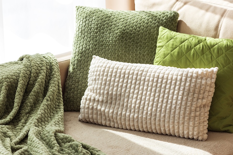 Three textured pillows—two green and one white—are arranged on a beige sofa next to a green throw blanket.