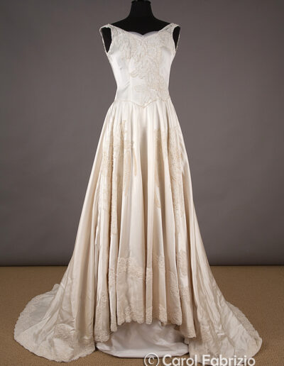 A vintage ivory satin wedding gown with lace detailing, a fitted bodice, wide straps, and a full skirt with a train displayed on a black dress form against a neutral backdrop.