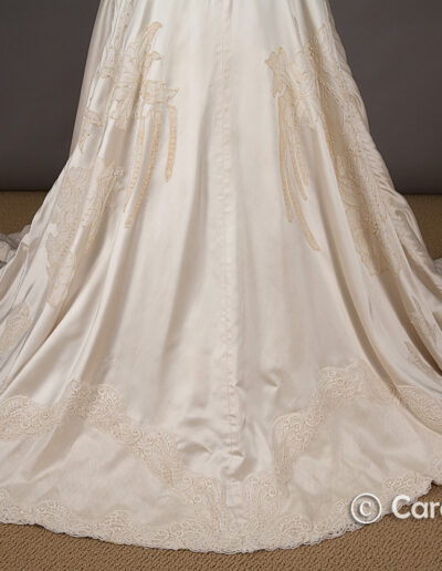 A close-up of the lower portion of an ivory lace wedding gown with a long train, displayed on a beige carpet against a gray background.