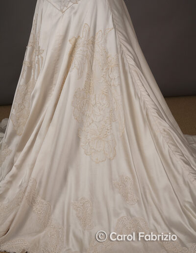 Cream-colored satin wedding dress with intricate lace embroidery along the hem and train, displayed against a neutral background.