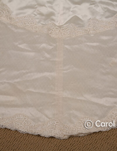 Close-up of the hem of a white satin garment with lace trim, displayed on a tan carpet. The watermark "© Carol Fabrizio" is visible in the lower right corner.