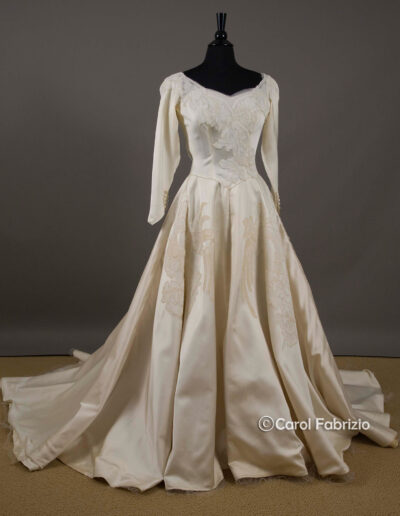 Vintage ivory satin wedding dress with long sleeves, off-shoulder neckline, embroidered bodice, and full skirt displayed on a black dress form against a plain background.