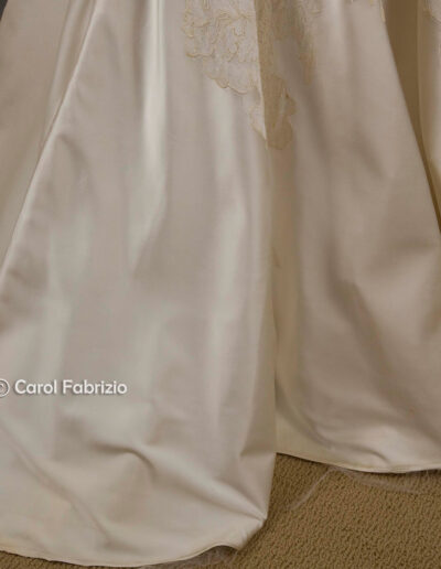 Close-up of the lower portion of a white satin wedding dress with lace appliqué details, touching a beige carpeted floor.