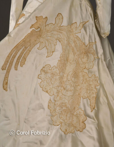 Close-up of an ivory satin fabric with an ornate appliqué floral lace pattern in beige tones; part of a vintage garment, possibly a dress or gown.