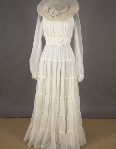 A long-sleeved, floor-length white dress with a tiered skirt and sheer fabric, displayed with a wide-brimmed matching hat adorned with floral details, set against a plain background.