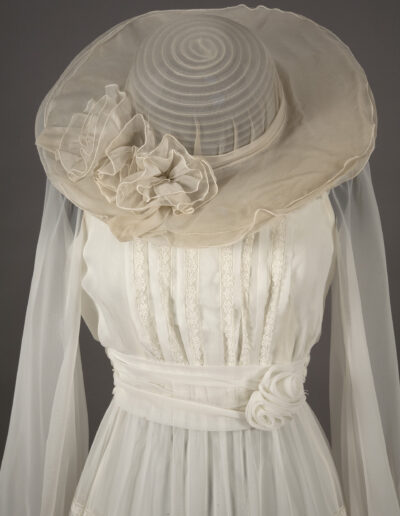 A vintage white lace dress with long sheer sleeves, a wide-brimmed translucent hat, and fabric flower details at the waist and hat.