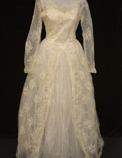 A vintage white lace wedding dress with long sleeves, a high neckline, and a full tulle skirt displayed on a mannequin against a black background.