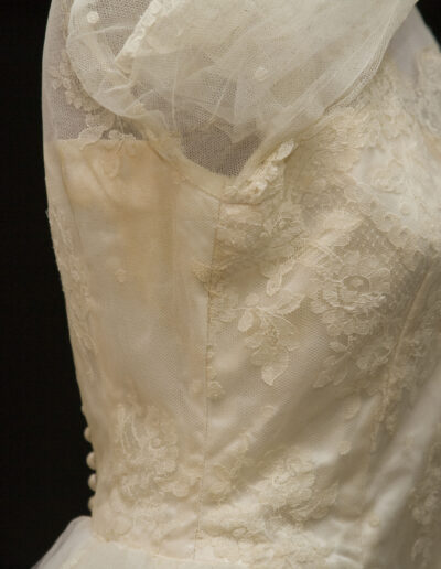 Close-up of a vintage white lace wedding dress with fitted bodice, floral patterns, and sheer tulle sleeves, displayed on a mannequin against a black background.