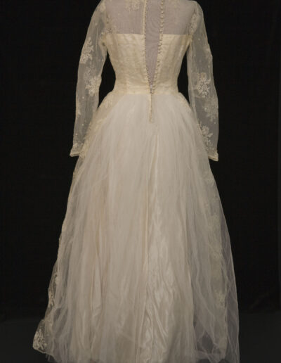 A vintage white wedding dress with lace long sleeves, sheer upper bodice, fabric-covered buttons down the back, and a full tulle skirt, displayed on a mannequin against a black background.