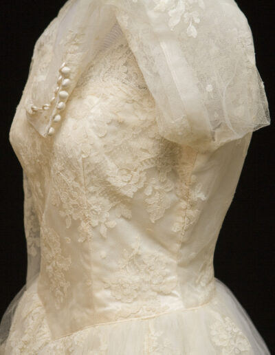 Ivory lace wedding dress with short sleeves, decorative buttons on the bodice, and a tulle overlay, shown on a mannequin against a black background.