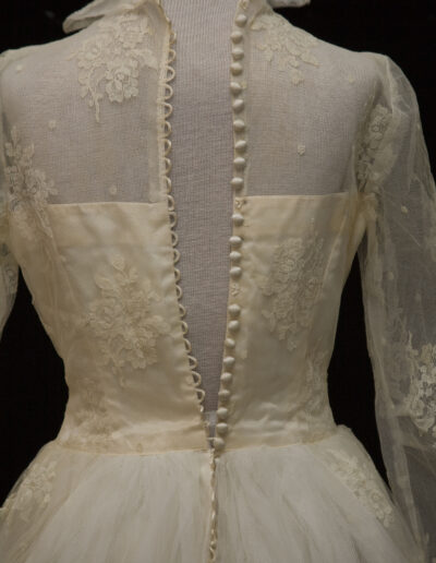 Close-up of the back of a vintage white lace dress with floral embroidery, sheer fabric, fabric-covered buttons, and a high collar on a mannequin.