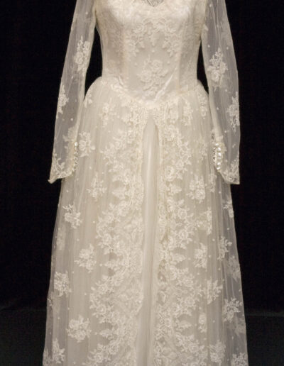 Long-sleeved white lace wedding dress with a high neck, sheer overlay, floral patterns, and button details on the cuffs, displayed on a mannequin against a black background.