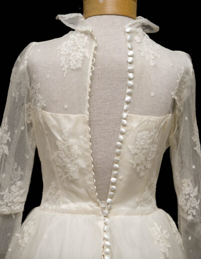 Back view of a vintage white lace wedding dress on a mannequin, featuring long sleeves, floral embroidery, and a row of white buttons down the back.