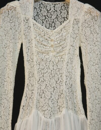 Long-sleeved white lace dress with buttons and lace-up detailing on the bodice, displayed on a wooden hanger against a black background.