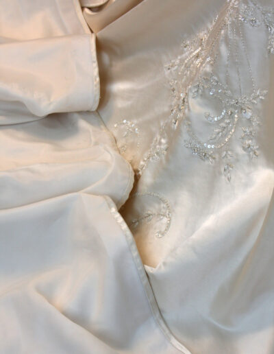 Close-up of ivory satin fabric with delicate embroidery and beadwork, possibly part of a wedding dress or formal gown.