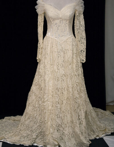 A vintage ivory lace long-sleeve wedding gown with a fitted bodice, puffed shoulders, and a long train, displayed on a mannequin against a black background.