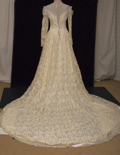 An off-white, long-sleeved lace wedding dress with a fitted bodice, buttoned back, and a long, flowing train displayed on a mannequin.