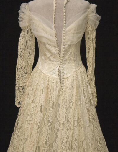 A vintage ivory lace dress with long sleeves, off-the-shoulder tulle detail, and a row of buttons down the back, displayed on a mannequin against a black background.