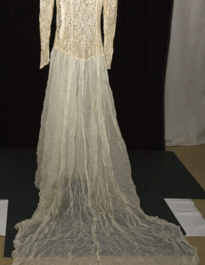 A long-sleeved, floor-length vintage lace wedding dress with a fitted bodice and a sheer, flowing train displayed against a black background.