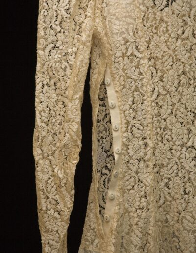 Close-up of a cream-colored lace garment with long sleeves and a row of white buttons along the side opening, set against a black background.