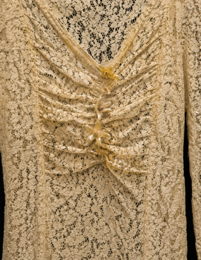 Close-up of a beige lace fabric with a ruched detail at the center, held together by a row of translucent buttons and small yellow fabric flowers.