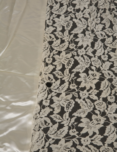 Close-up of a piece of sheer white lace fabric with a floral pattern, placed next to a shiny, smooth, off-white satin fabric.
