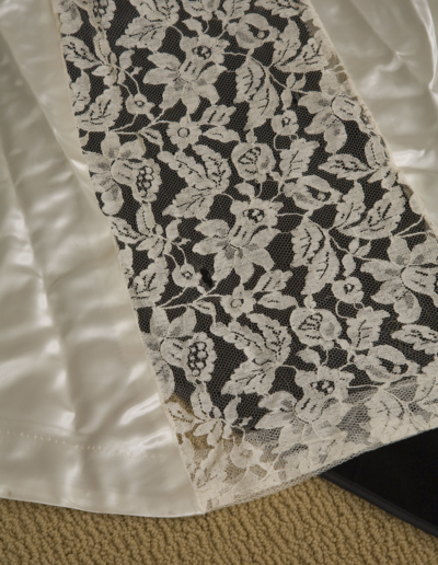 A close-up of a white lace fabric panel with a floral pattern laid over a shiny white satin material on a beige carpet.