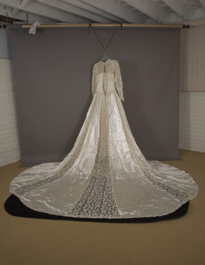 A long-sleeved vintage wedding dress with lace detailing and a floor-length train is displayed on a hanger against a gray backdrop in a room with white brick walls.