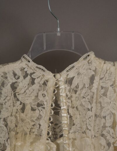 Close-up of a cream lace garment with floral patterns, decorative buttons, and a beaded neckline, displayed on a clear plastic hanger against a plain background.