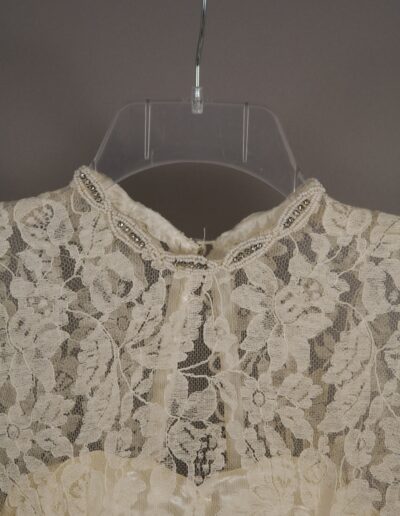 Close-up of a vintage cream lace garment on a clear plastic hanger, featuring a beaded neckline and floral lace pattern.