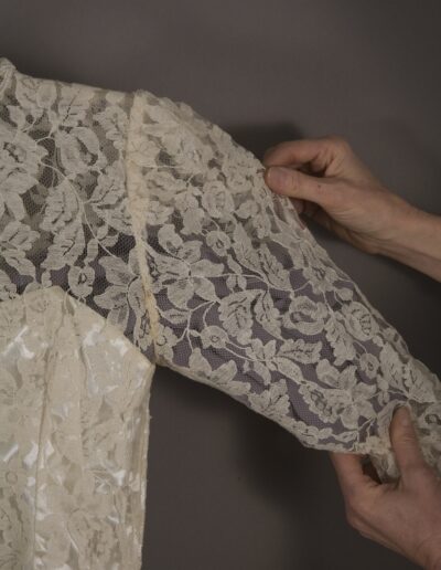 A person holds the lace sleeve of a cream-colored garment against a gray background, showing the floral lace pattern in detail.