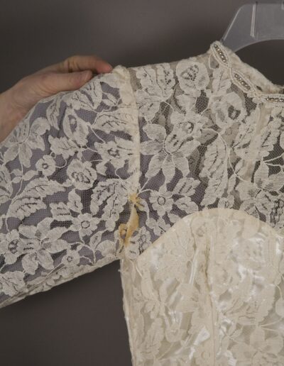 Close-up of a hand holding a vintage cream lace dress on a hanger, showing a tear and yellowed area under the arm of the sheer lace sleeve.