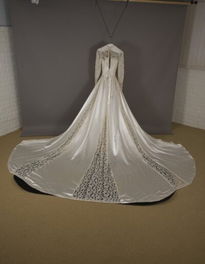 A long-sleeved white wedding gown with a long lace train is displayed on a mannequin against a gray backdrop in a room with white walls and beige carpet.