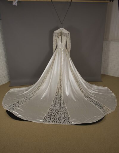 A long-sleeved, floor-length white satin wedding dress with lace panels, displayed on a mannequin against a gray backdrop.