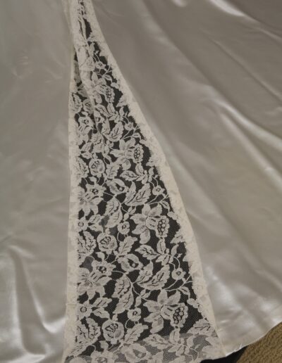 Close-up of a white satin fabric with a triangular lace panel featuring a floral pattern sewn into it. The fabric is spread out on a flat surface.