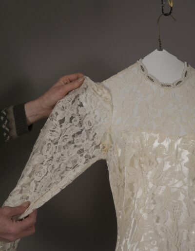 Person holding the lace sleeve of an off-white vintage dress on a hanger, showing the detailed lace pattern and beaded neckline.