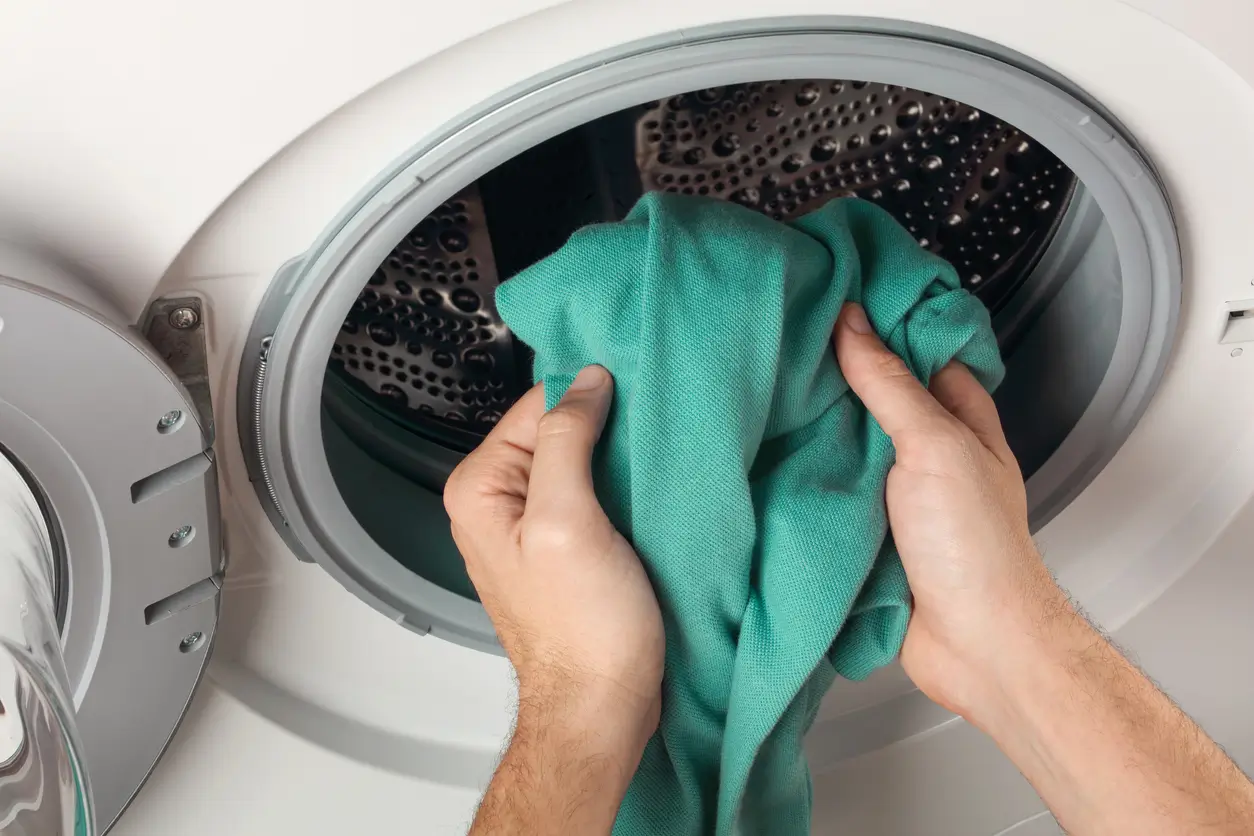Hands placing a green garment into the open drum of a front-loading washing machine. Hands placing a green garment into the open drum of a front-loading washing machine.